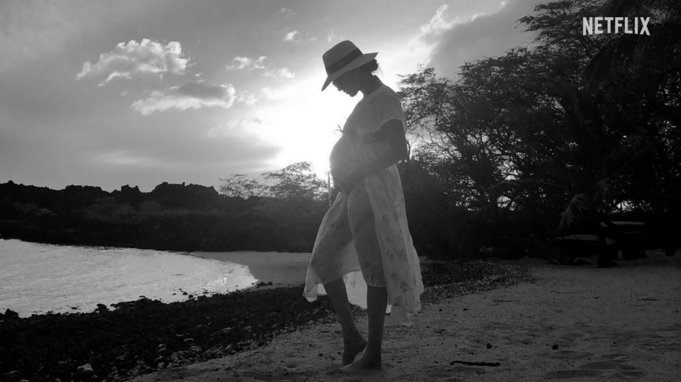 Meghan holds her pregnant stomach while standing on a beach.