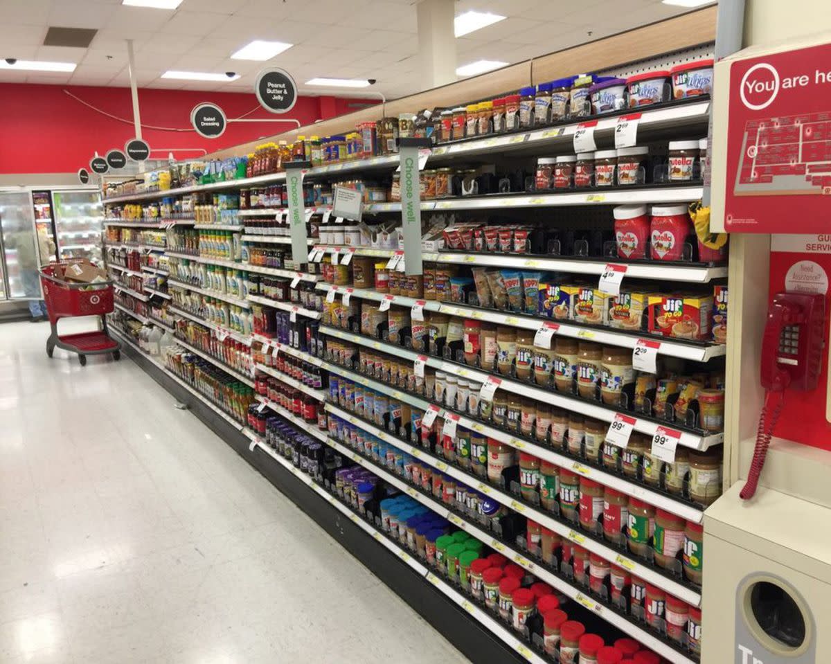 An aisle in the grocery section of a Target store, peanut butter, spreads in the right foreground, salad dressings and other condiments in the background left, with a red shopping cart, freezers lining the back area, a person retrieving items from a