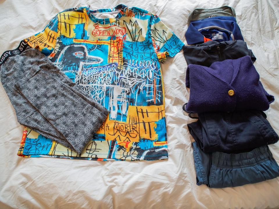 From left to right: folded gray leggings with a black pattern on top of a blue and yellow t shirt and  and a row of two rolled-up shirts and two fold-up long sleeve layers that are gray, black, navy blue Folded teal pants, and folded black pants on a white sheet