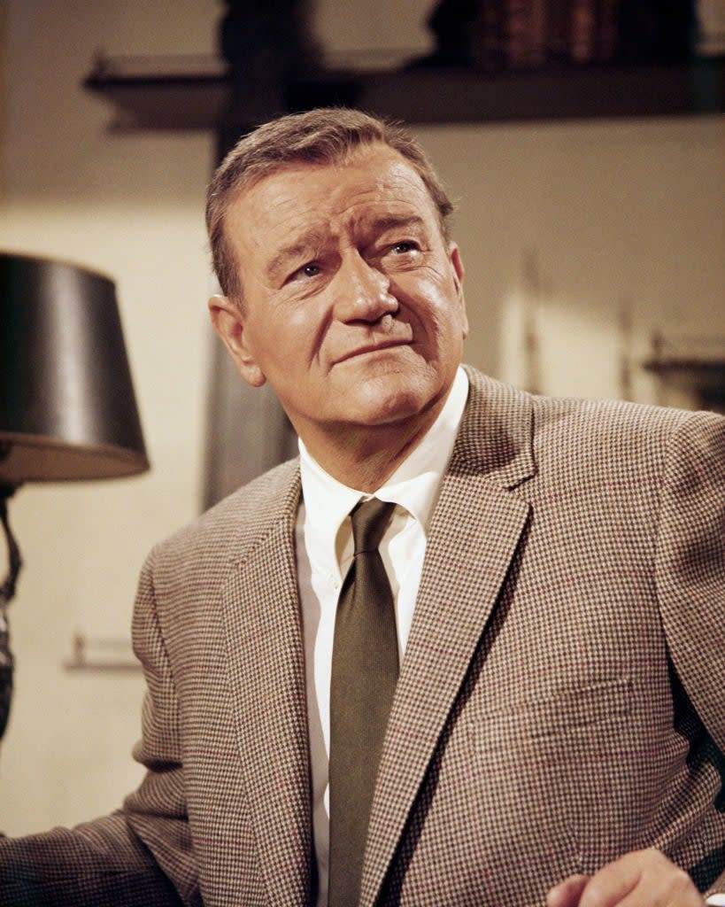John Wayne in a tweed suit, smiling slightly, in a vintage indoor setting