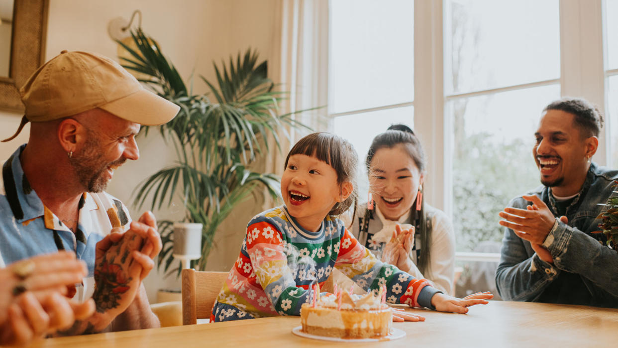  young child sits at a dining table surrounded by adults who laugh and clap for her as she blows out birthday candles on a cake. 