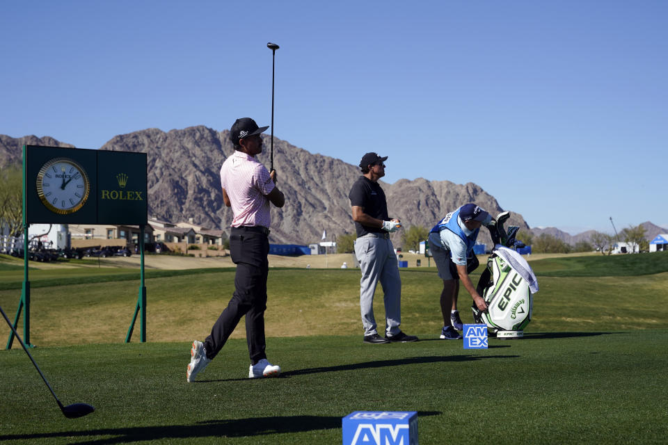 Rickie Folwer, left, hits from the first tee as Phil Mickelson, center, watches during the first round of The American Express golf tournament on the Nicklaus Tournament Course at PGA West, Thursday, Jan. 21, 2021, in La Quinta, Calif. (AP Photo/Marcio Jose Sanchez)