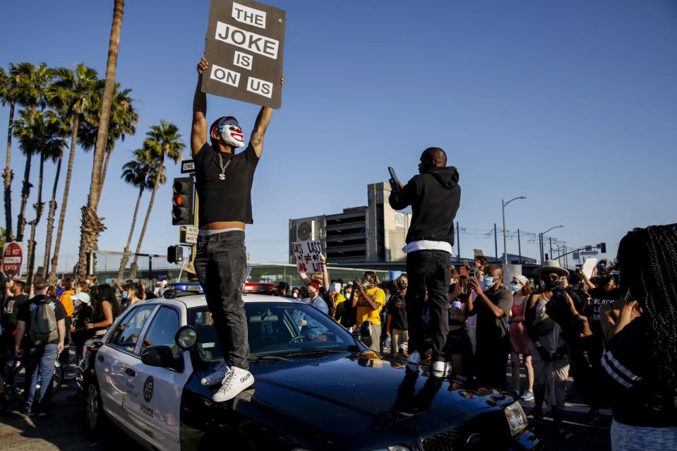 Two people, one holding a sign reading "The joke is on us," stand on a police vehicle during a Black Lives Matter protest