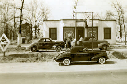 Late-1930s:  Many of stock car racing’s early stars learned their driving trade as “bootleggers” hauling illegal liquor.  Among them were Smokey Purser, Lloyd Seay, Roy Hall and Joe Littlejohn, whose racecar (No. 7) is seen outside the “Little Joe’s” tavern.  Note how Littlejohn’s racer did not look much different than the street cars in the photo.