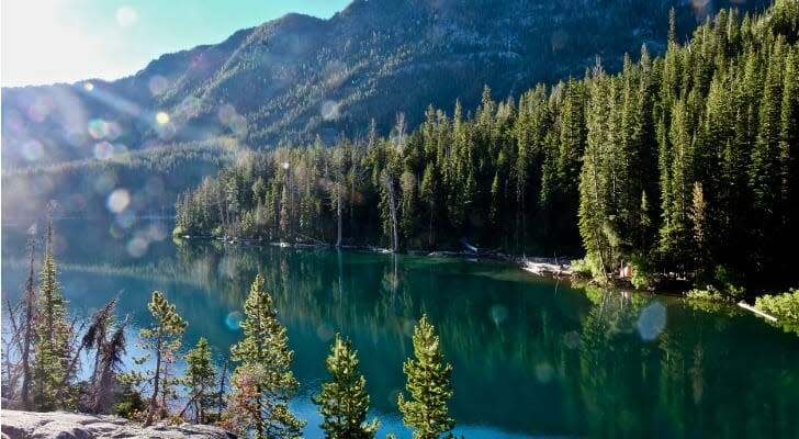 Image shows the sun shining brightly over a clear lake surrounded by evergreen trees at the foot of a mountain in the Pacific Northwest. In this study, SmartAsset analyzed various data sources to find the best places for outdoor enthusiasts to live and work.