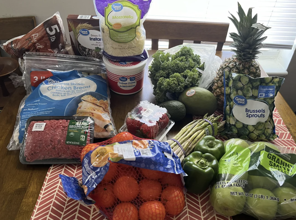 Various groceries on a table include chicken breast, mozzarella cheese, yogurt, pineapple, kale, mango, ground beef, Brussels sprouts, asparagus, bell peppers, apples, and a bag of oranges