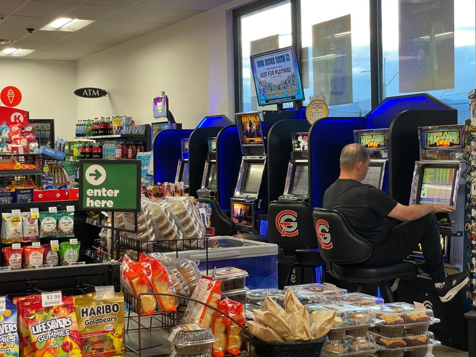 A man sitting at a line of casino machines inside a gas station with a table of snacks behind him.