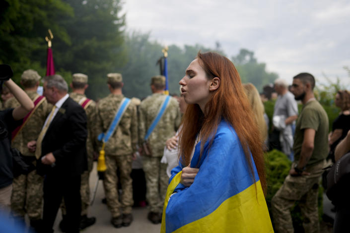 A woman wrapped in a Ukrainian flag attends the funeral of activist and soldier Roman Ratushnyi in Kyiv, Ukraine, Saturday, June 18, 2022. Ratushnyi died in a battle near Izyum, where Russian and Ukrainian troops are fighting for control of the area. (AP Photo/Natacha Pisarenko)