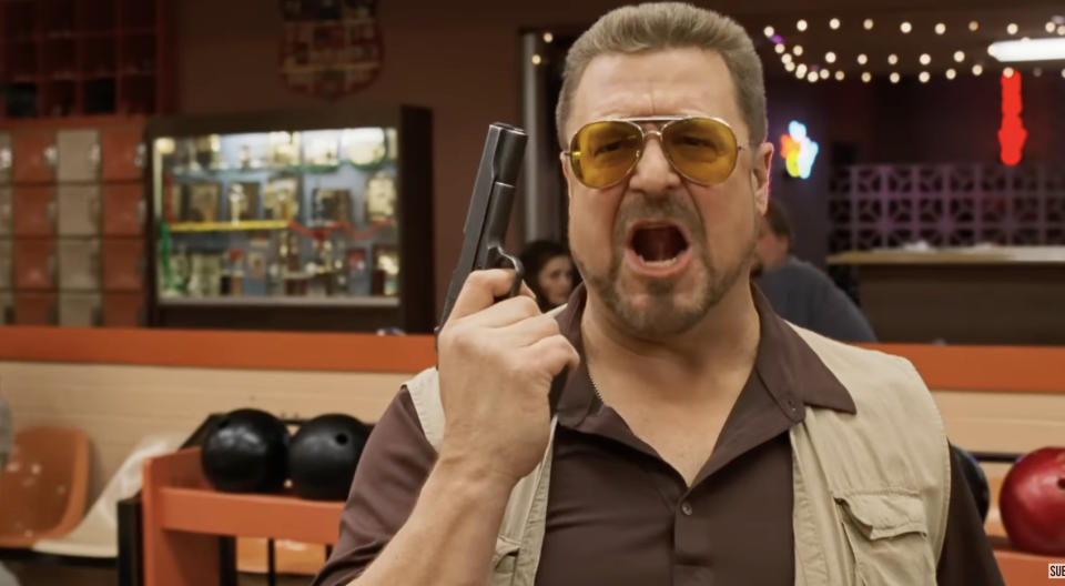 Man wearing aviator sunglasses and a beige vest, loudly speaking while holding a handgun at a bowling alley, from The Big Lebowski