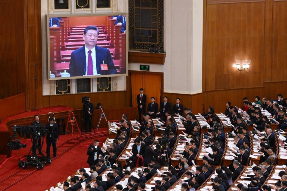 Xi Jinping on a TV screen overlooking a large room full of seated delegates.