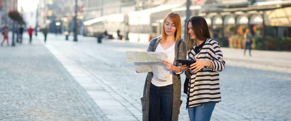 Two young women walk on unfamiliar city. Girlfriends go sightseeing. In hands women have a card. They are surrounded with fine architecture. Tourists have a good mood.