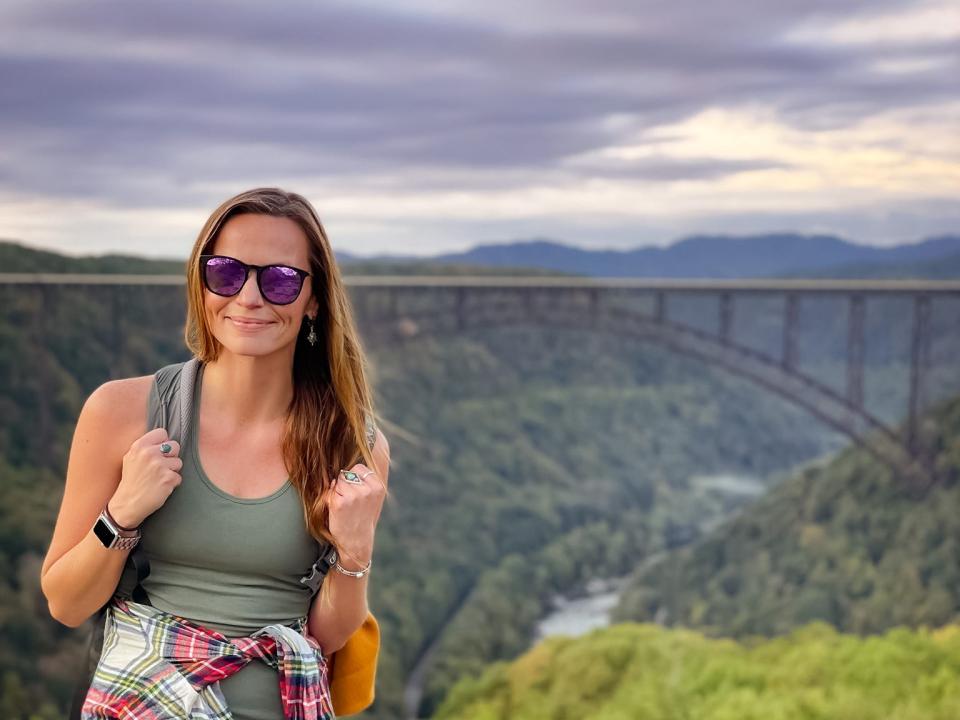 Emily, wearing sunglasses, black leggings, a green tank top, and a flannel around her waist, stands on rocks in New River Gorge National Park, with a bridge behind her.