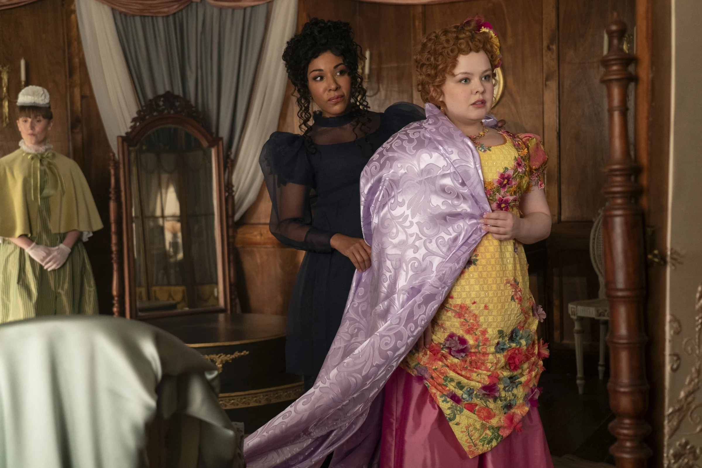 Nicola Coughlan, in a floral gown with a purple shawl, stands in front of an ornate mirror. Another woman in a dark dress assists her. A third woman watches in the background