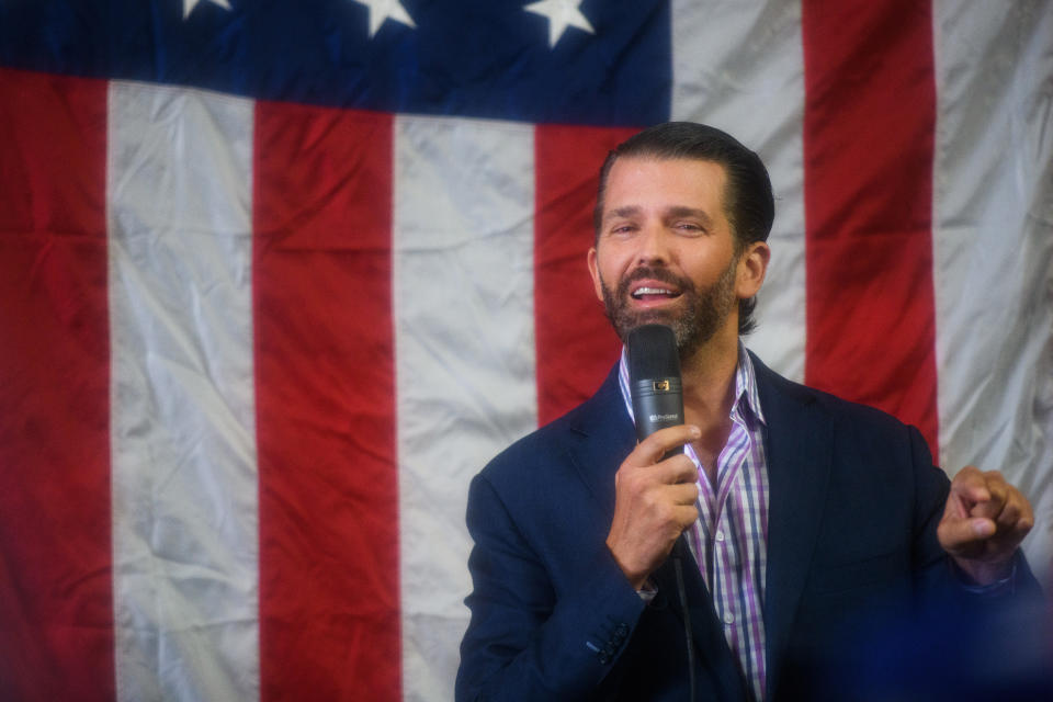 Donald Trump Jr., holding a microphone, speaks in front of an American flag.