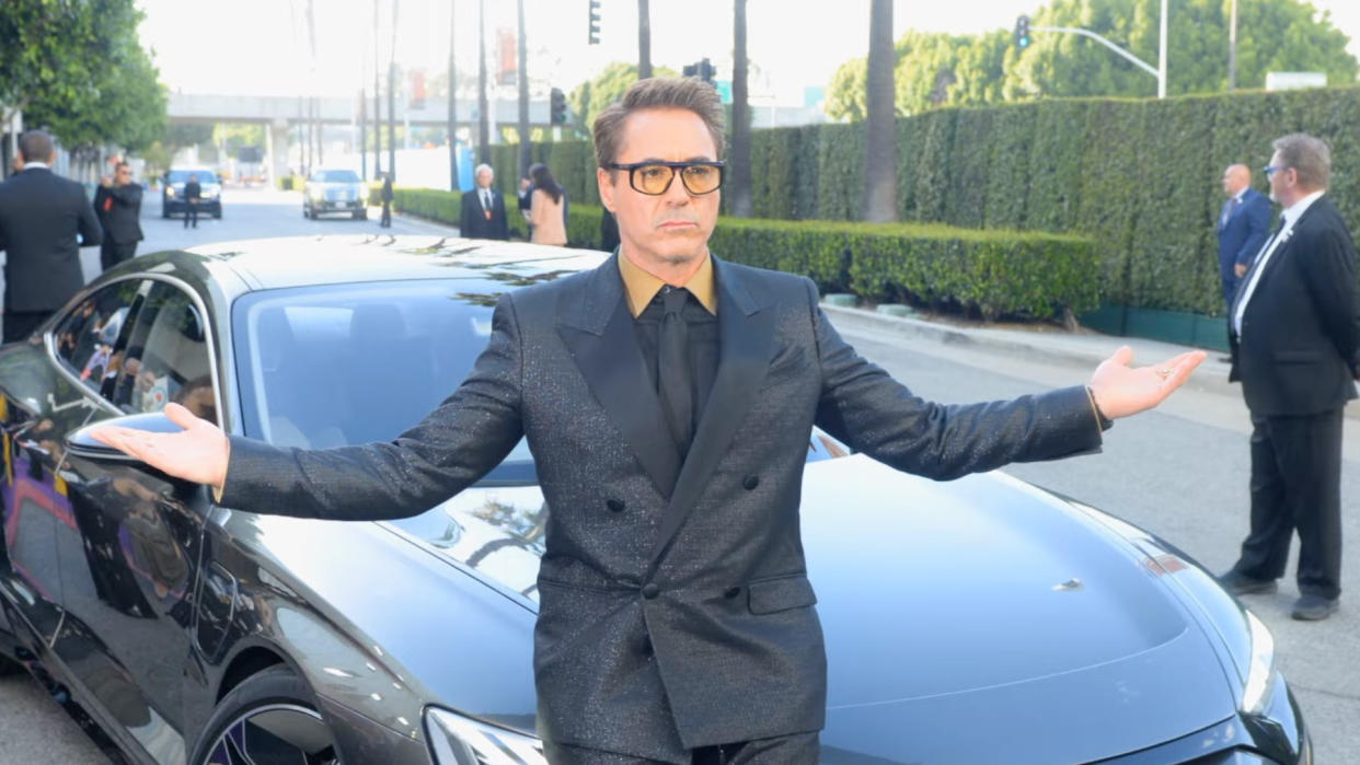  Robert Downey Jr., wearing a black suit and sunglasses, stands in front of a car with his arms in the air, in a trailer for Downey's Dream Cars shown at the WBD Upfronts 2023 