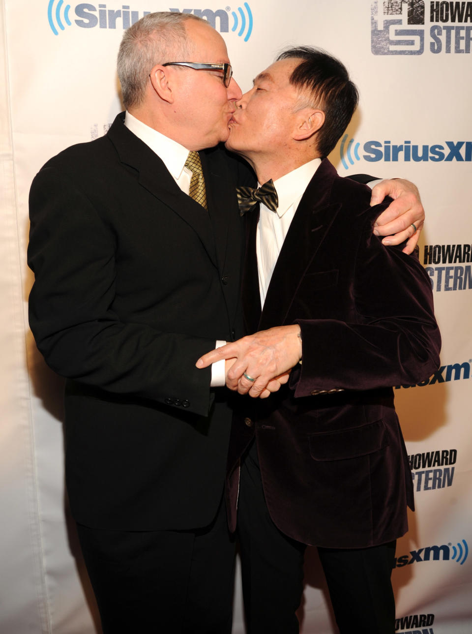 George Takei and Brad Altman share a kiss while embracing, standing in front of a step-and-repeat banner featuring SiriusXM and Howard Stern logos