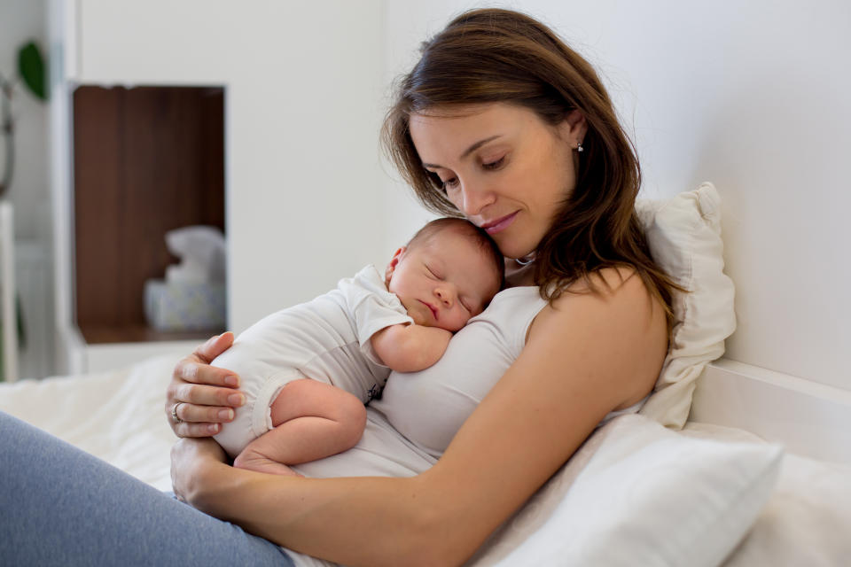 Young mother lying in bed with her newborn baby boy, holding him in her arms and smiling from happiness