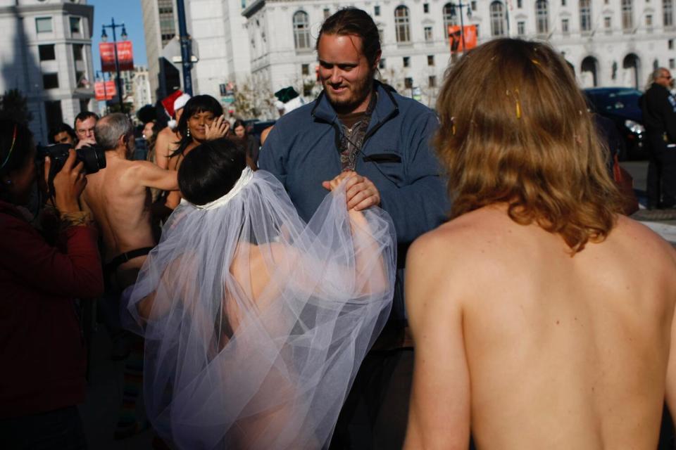 A man grasps the hand of a bride wearing only a head veil at a nude wedding.