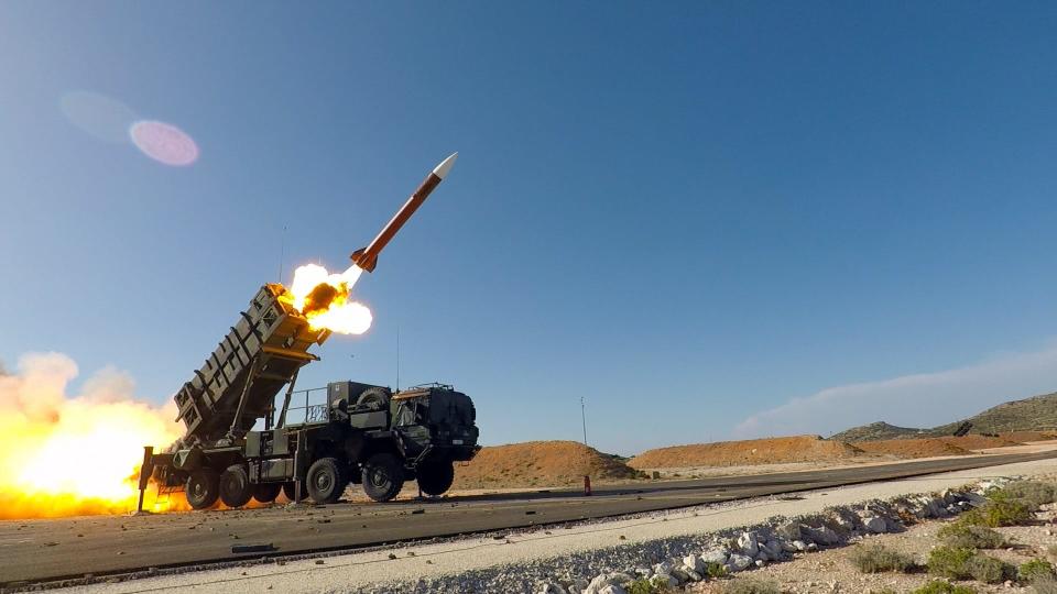 A Patriot air defense system fires an interceptor missile in Greece, with flames coming from the launcher.