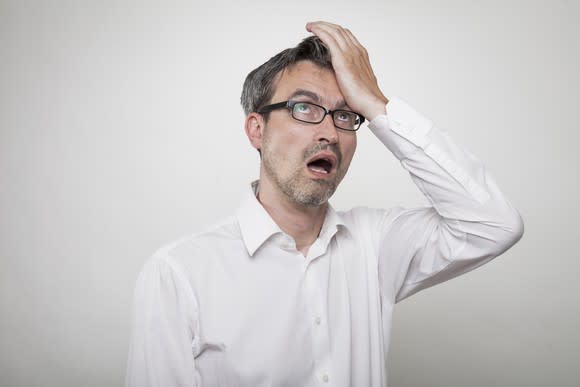 A man with glasses and a white shirt holds his palm to his forehead