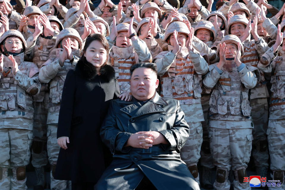 Kim Jong Un, seated, with his daughter, Kim Ju Ae, standing at his shoulder, with rows of troops in camouflage behind them raising their hands to applaud.
