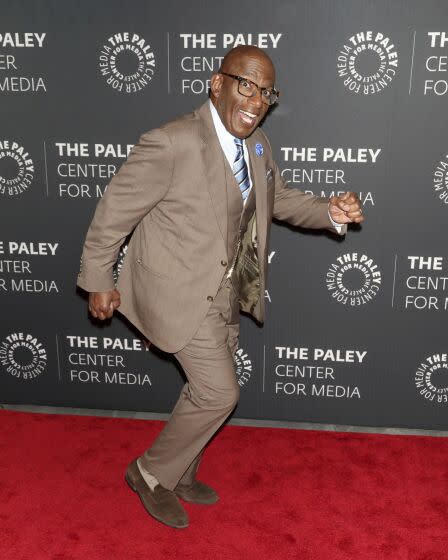 A man in a three-piece suit smiles and goofs around on a red carpet