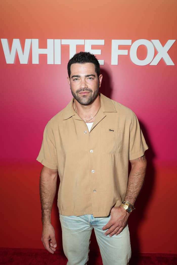 Jesse Metcalfe at the 'White Fox' event. He is wearing a beige button-up shirt and light blue jeans, posing in front of a red and pink gradient backdrop