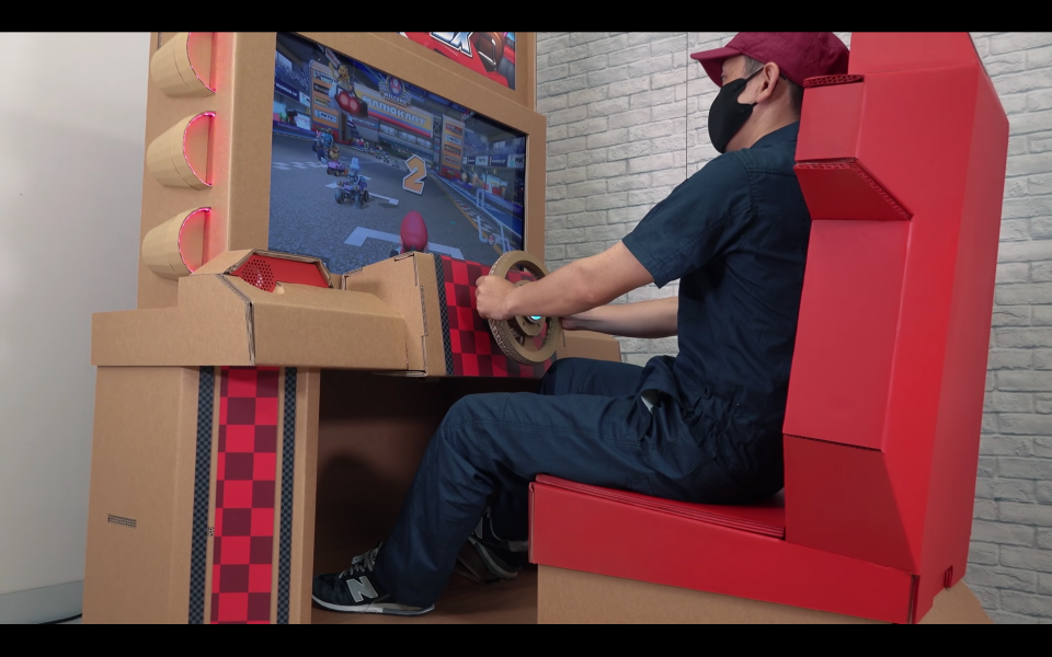 A master cardboard craftsperson playing a Mario Kart 8 arcade machine made out of cardboard pieces and relatively simple electronic components.