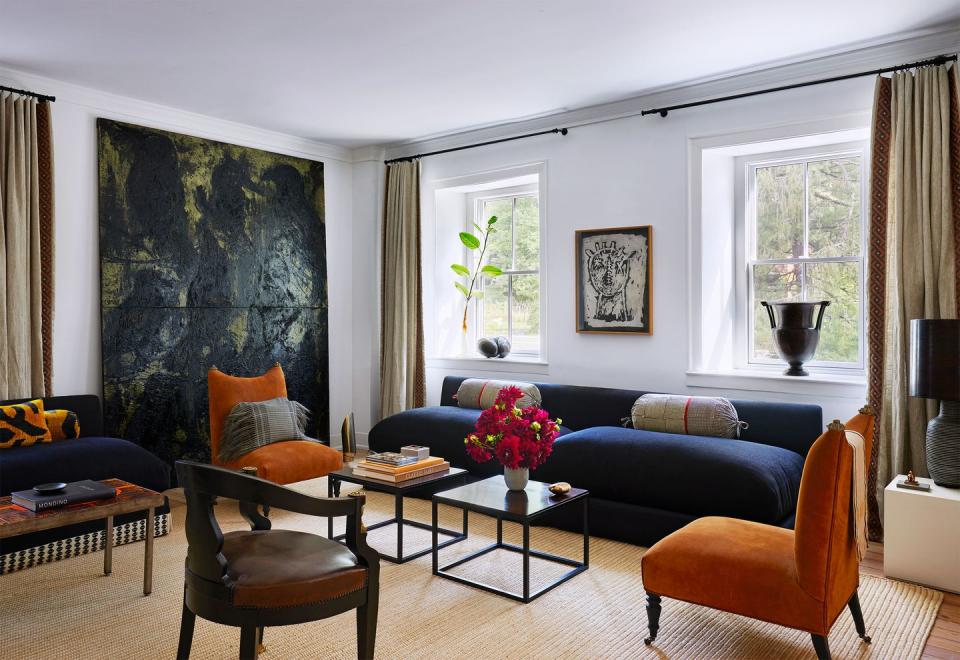 in a living room are two dark blue low backed sofas, two armless chairs in a deep orange velvet, twin small cocktail tables, an ebony chair, a steel topped cocktail table, and a large artwork propped against one wall
