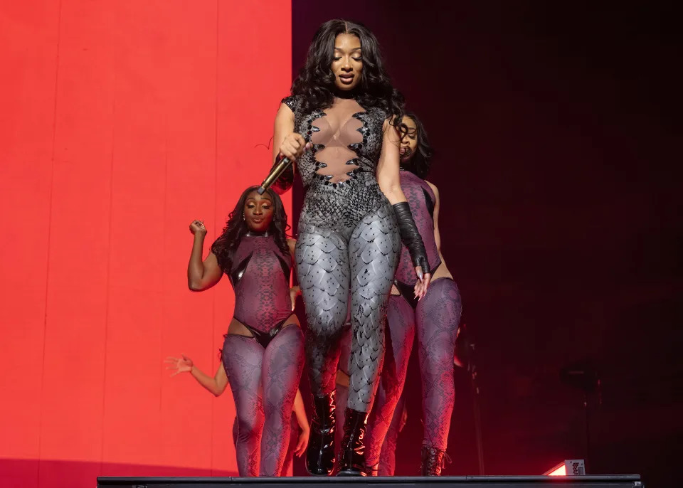 Megan Thee Stallion performs on stage in a sheer, form-fitting outfit with intricate patterns, accompanied by three backup dancers in similar outfits