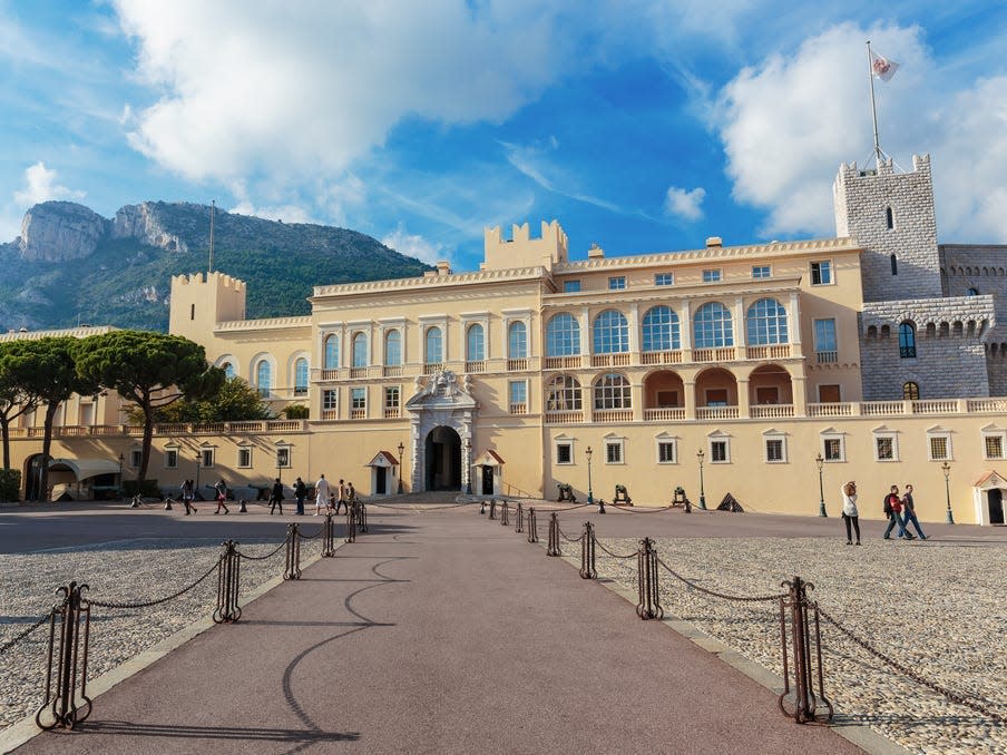 The Prince's Palace of Monaco. There are mountains behind the building and people walking around the grounds.