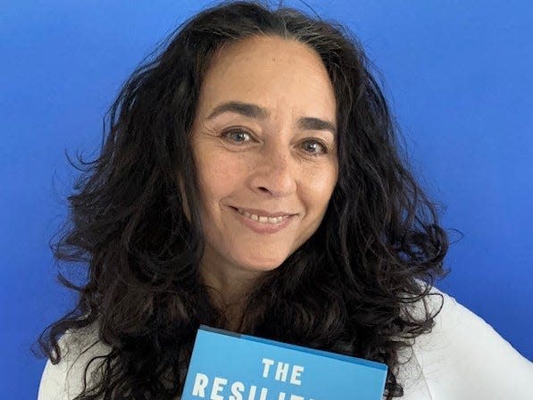 Soraya Chemaly standing against a blue background holding a copy of her book "The Resilience Myth: New Thinking on Grit, Strength, and Growth After Trauma."