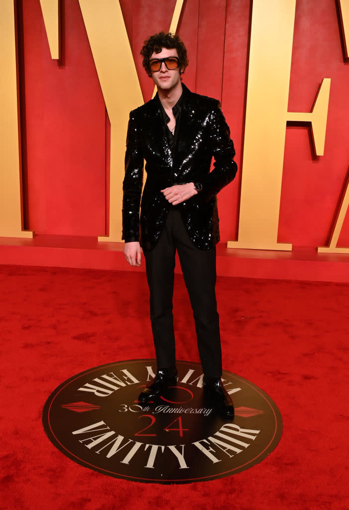 Man on red carpet wearing a sequined black suit and curly hair, standing on Vanity Fair logo