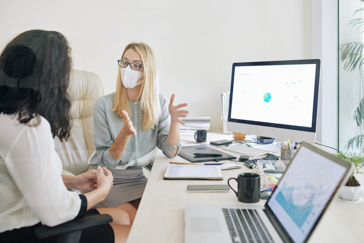 Young businesswoman in glasses and medical mask actively gesturing when explaining idea to collesgue