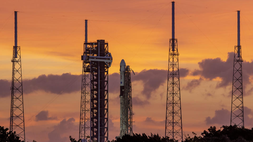     Eine weiße Rakete steht auf einer Startrampe, umgeben von vier hohen Blitztürmen vor einem orangefarbenen Himmel. 