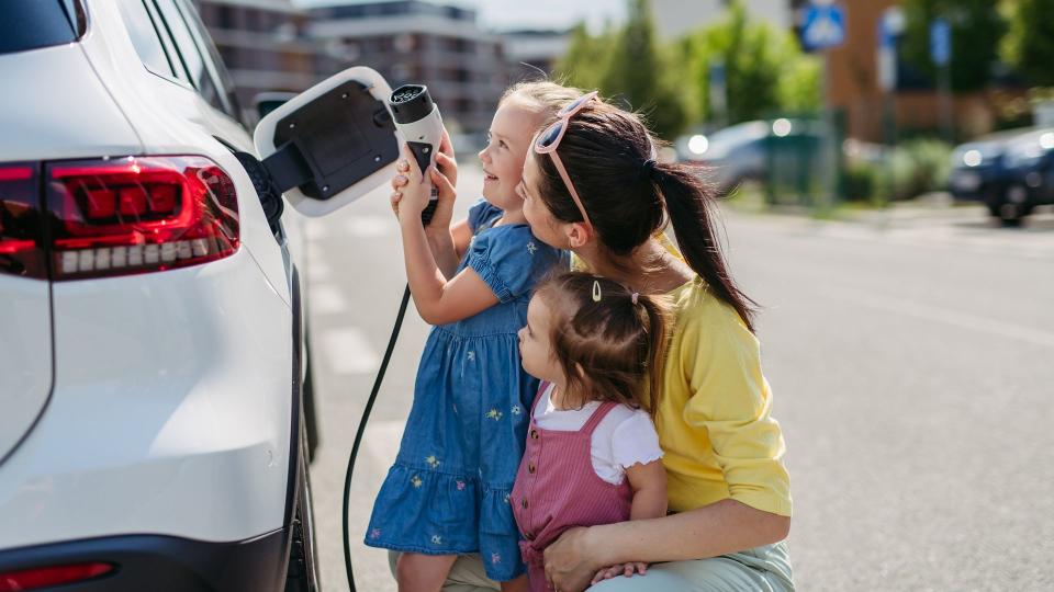 Mother and her little daughters charging their electric car on the street. Electric vehicle with charger in charging port. Cute girl helping to plug in charger.