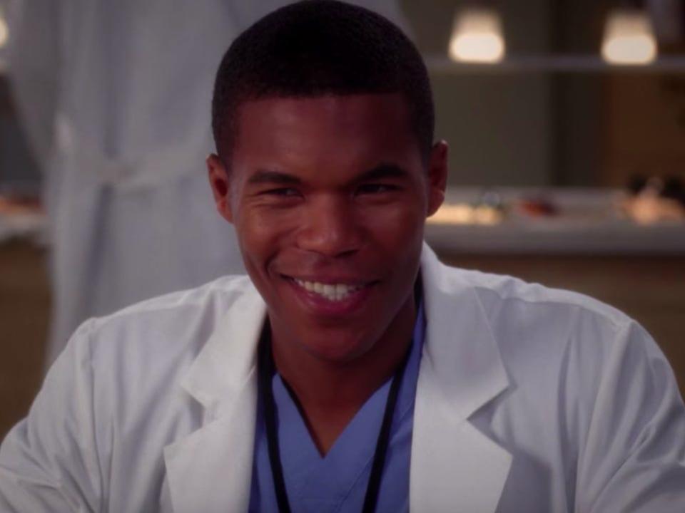 Gaius Charles as Shane Ross on Greys Anatomy wearing blue scrubs and a white coat