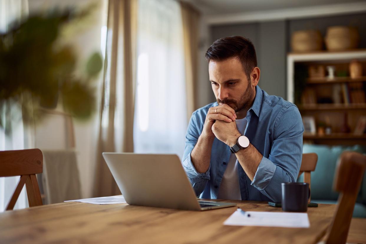 A deeply focused male freelance writer reading and revising his latest work on a laptop with his hands connected on his chin while sitting at a desk at home.