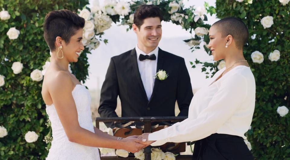 Two women dressed in white and getting married with male officiant.