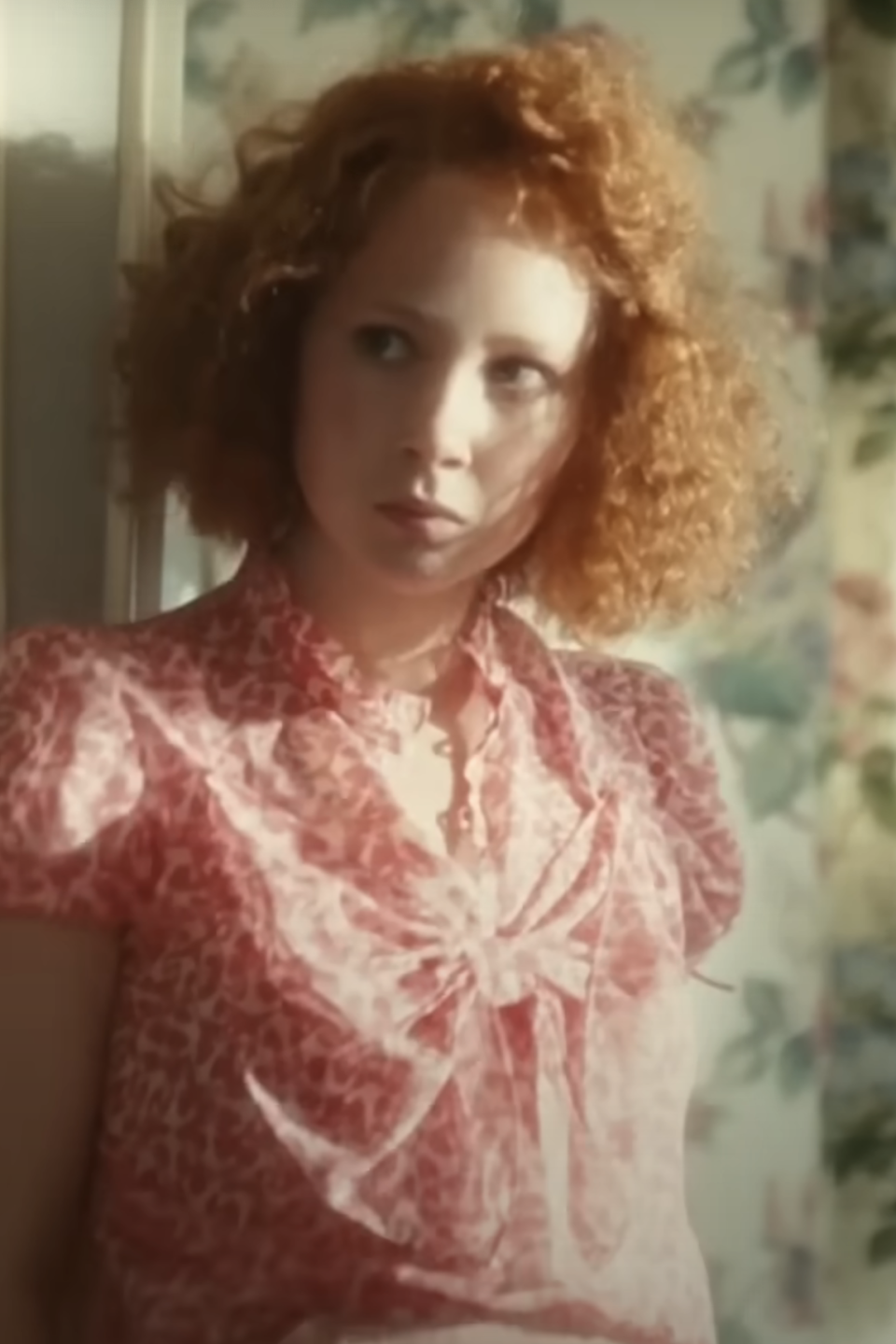 A young Juno Temple with curly red hair standing in a scene from 