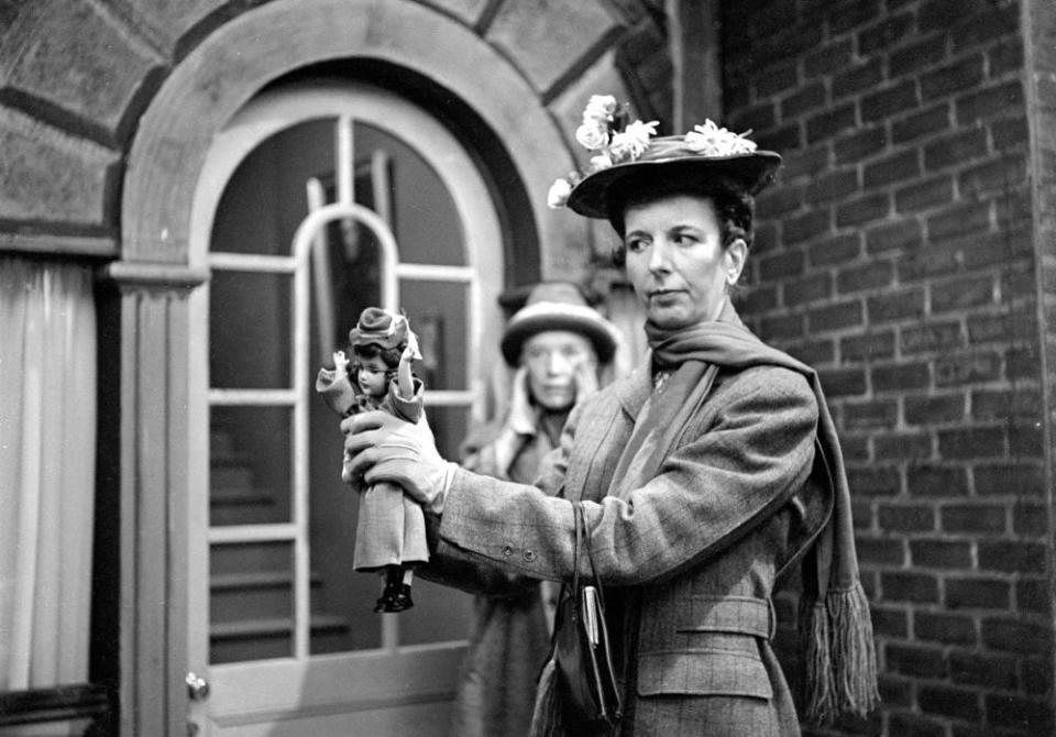 Two women in period attire stand outside a building. One holds a small puppet resembling the other woman