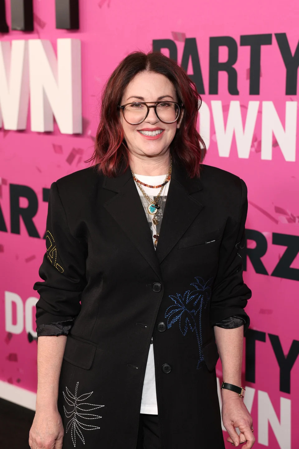 Megan Mullally attends a red carpet event wearing a black blazer with embroidered designs, layered necklaces, and glasses, smiling against a pink background