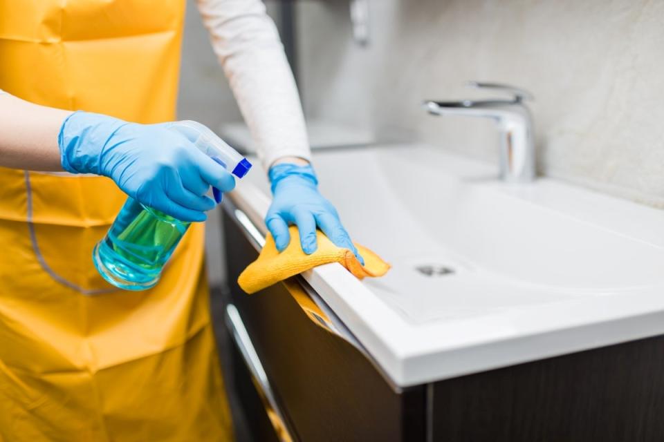 Woman wearing gloves and a yellow apron spraying and wiping a bathroom sink.