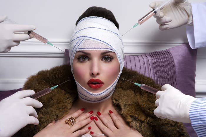 A woman is wrapped in bandages and surrounded by plastic surgeons who are holding injectables for cosmetic enhancements