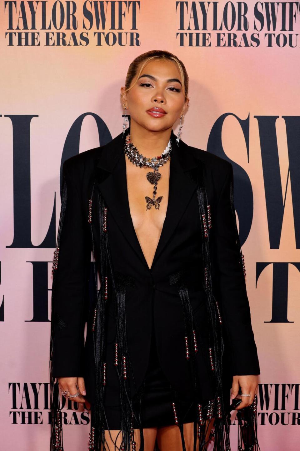 Hayley Kiyoko stands with hair slicked back in a black skirt and jacket with a necklace and no shirt