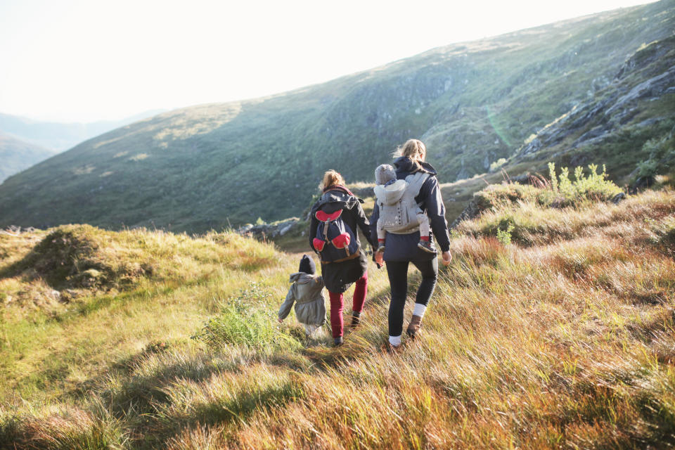 A family of four, with two children in backpacks, hikes through a grassy field in a mountainous area