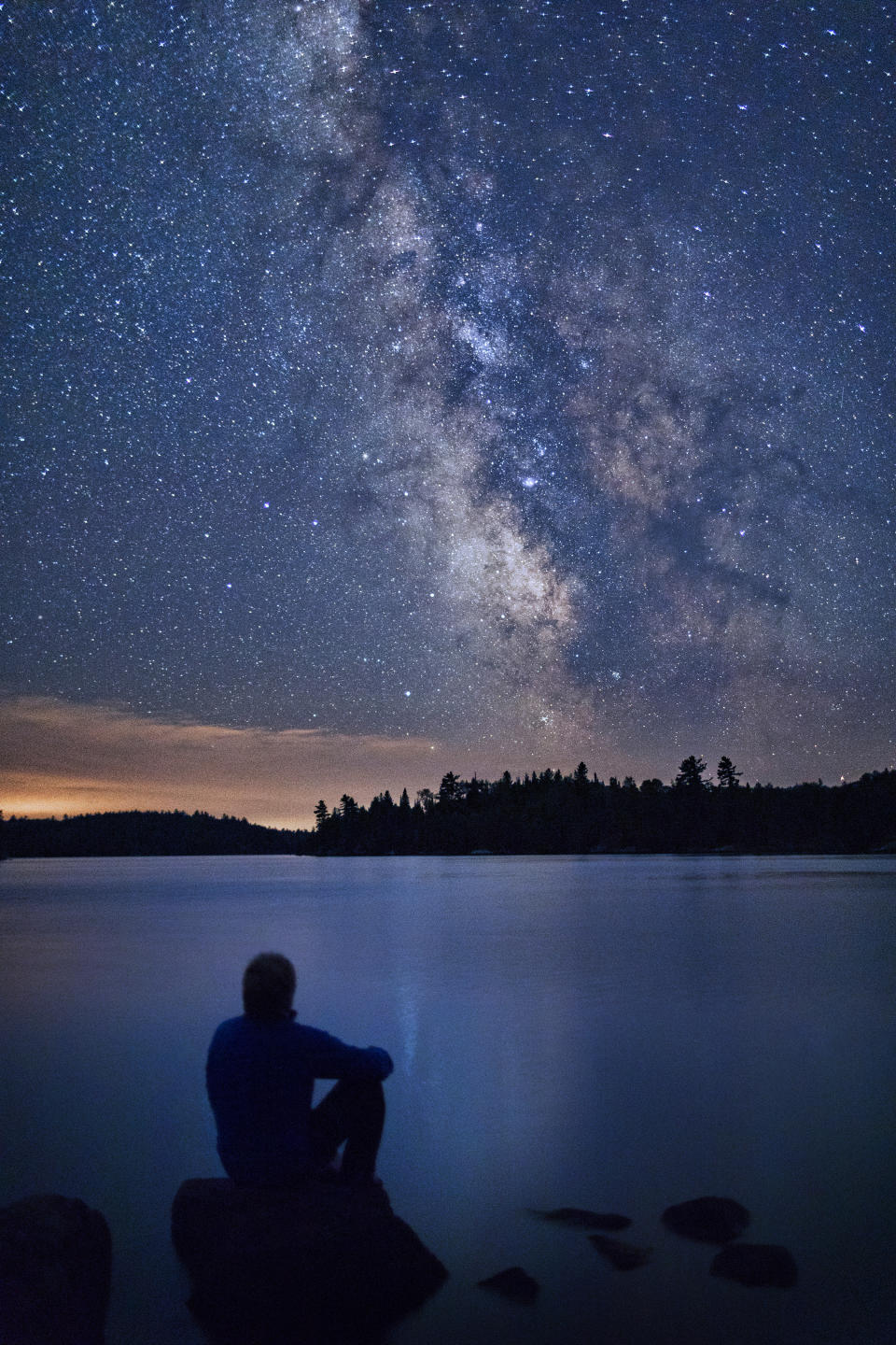 A man sits on a large boulder while looking out onto the dark and starry skies of Voyageurs national park.