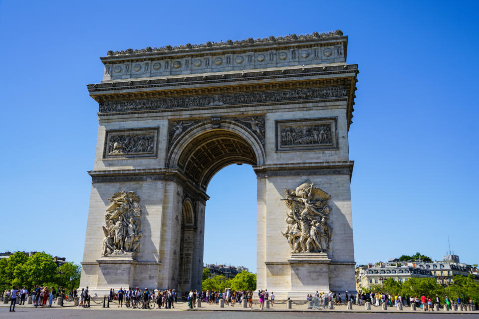 A day view of Arc de Triomphe de l'Étoile, one of the most famous monuments in Paris, France. The monument is in the sunlight and in the foreground of a clear summer sky. It is a popular tourist attraction. A number of people are visible at the base of the arc.