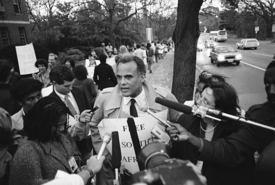 This black and white photo shows a man holding up a sign at a protest as reporter surround him.