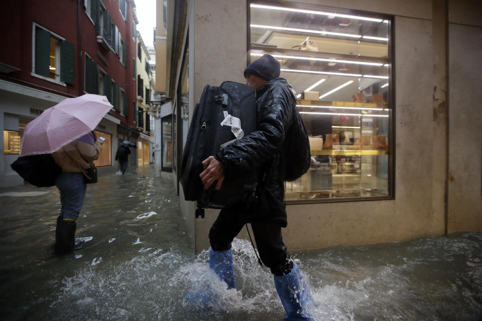 FILE - In this Friday, Nov. 15, 2019 file photo, a man holds his luggage as he wades his way through water in Venice, Italy. Venice's hoteliers association estimates that the city's hotels suffered about 30 million euros ($34 million) worth of structural damage during November's floods. The overall losses though are higher when the lower revenues that local hotels have reported in the wake of the surging high tides are added in. (AP Photo/Luca Bruno, File)